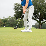 A male golfer in a blue polo shirt, white pants, and white golf shoes is mid-swing on a golf green, with a blurred white golf ball in the foreground and trees in the background.