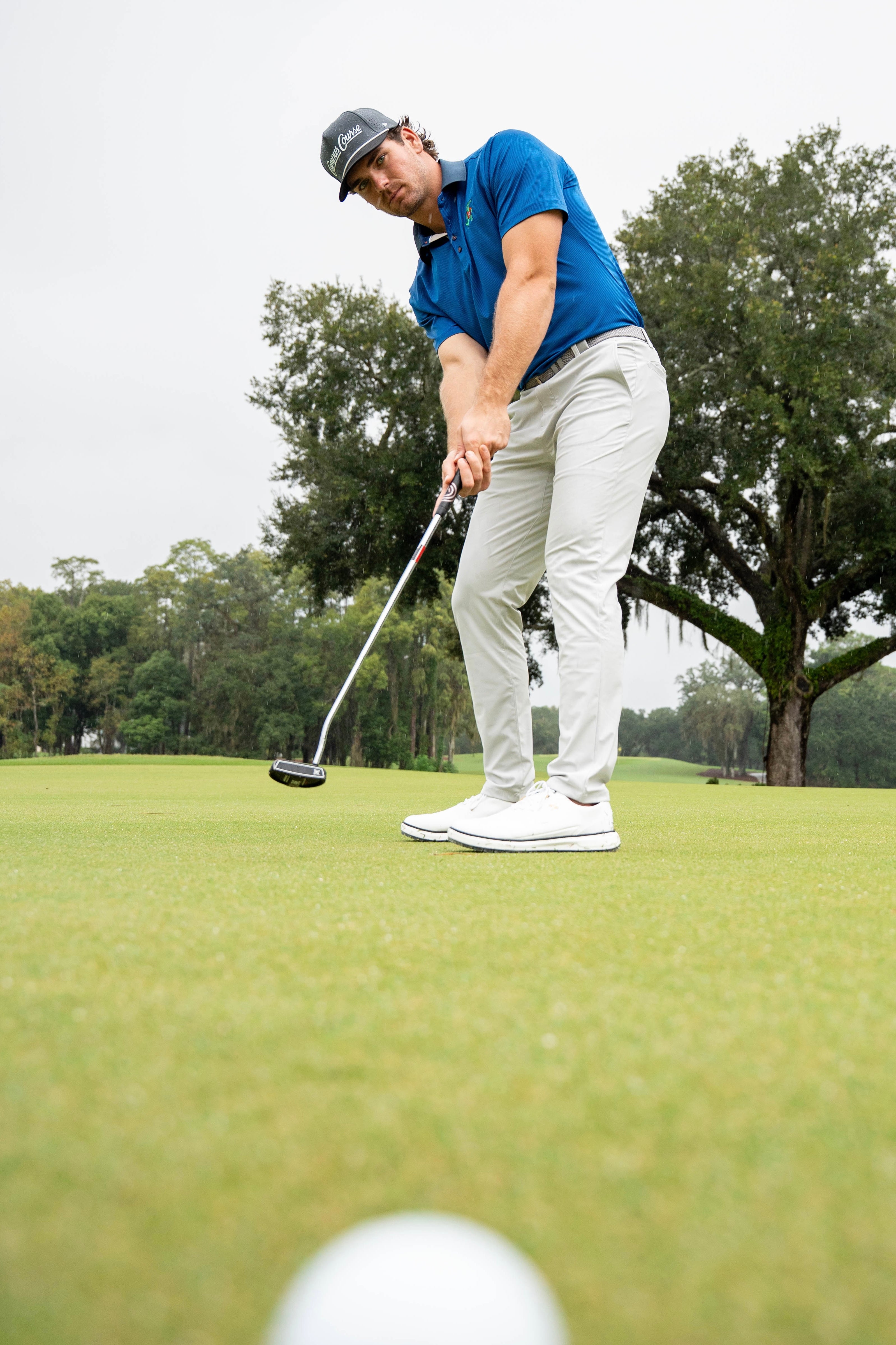 A male golfer in a blue polo shirt, white pants, and white golf shoes is mid-swing on a golf green, with a blurred white golf ball in the foreground and trees in the background.