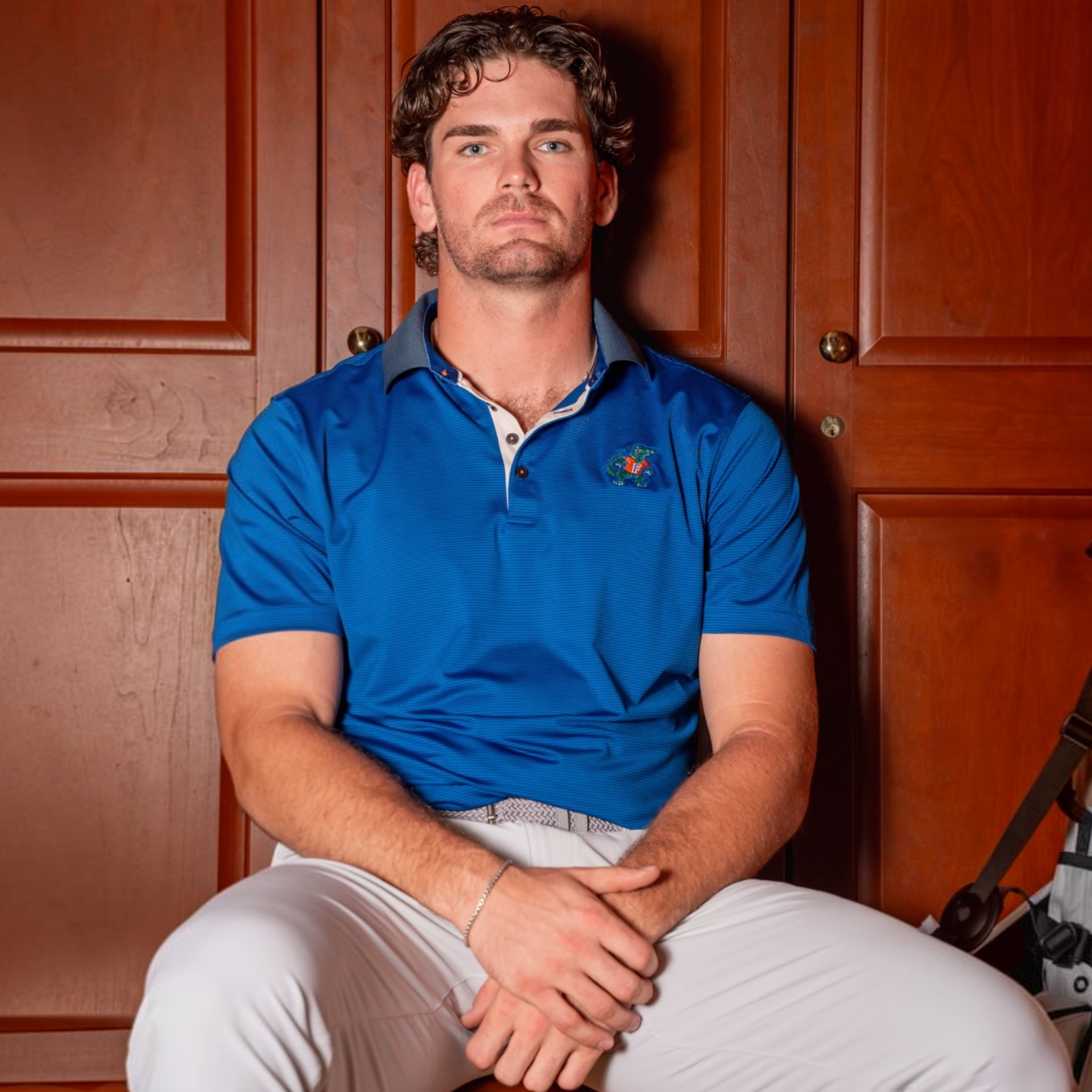 A man wearing a royal blue polo shirt with a small embroidered logo on the left chest, paired with white pants, sitting in front of warm brown wooden doors.