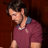 A man wearing a garnet polo shirt with a charcoal collar and a gold 'FSU' logo on the chest, photographed in a dimly lit indoor setting with dark wood paneling.
