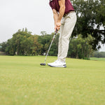 A man on a golf course putting green, wearing a garnet polo shirt, beige pants, white golf shoes, and a dark cap, with trees and an overcast sky in the background.