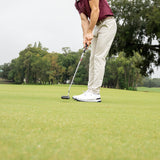 A man on a golf course putting green, wearing a garnet polo shirt, beige pants, white golf shoes, and a dark cap, with trees and an overcast sky in the background.