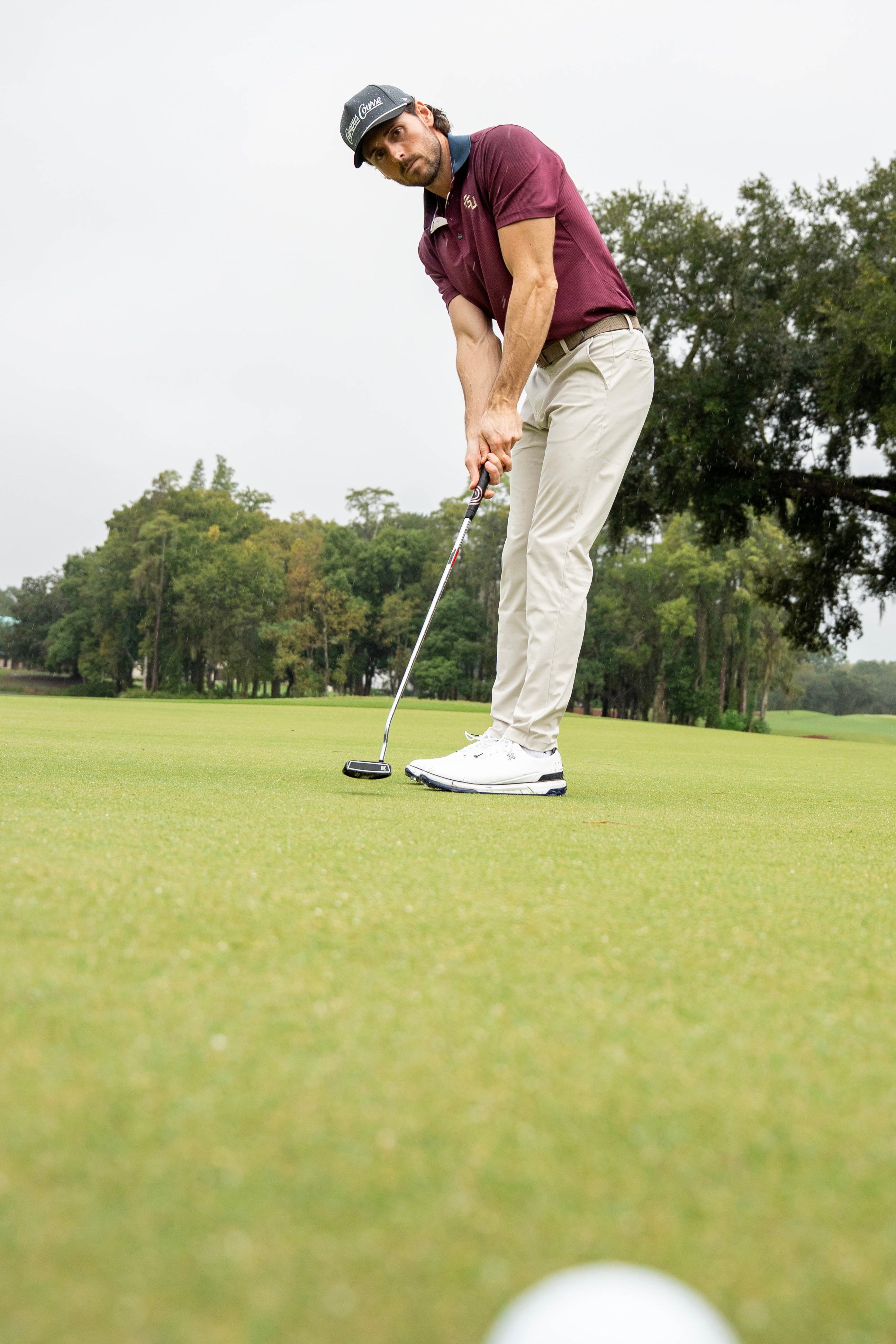 A man on a golf course putting green, wearing a garnet polo shirt, beige pants, white golf shoes, and a dark cap, with trees and an overcast sky in the background.