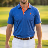 A man wearing a blue UF polo shirt with an orange collar and a small orange 'F' logo on the chest, paired with white shorts, standing on a golf course at sunset.