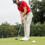 A man in a red polo shirt, beige pants, and a black cap is shown from behind as he prepares to putt a golf ball on a green course with trees in the background.