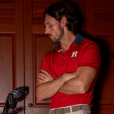 A man wearing a red short-sleeve polo shirt with a grey collar and a white letter 'R' logo on the chest, standing in front of dark wood doors with his arms crossed.